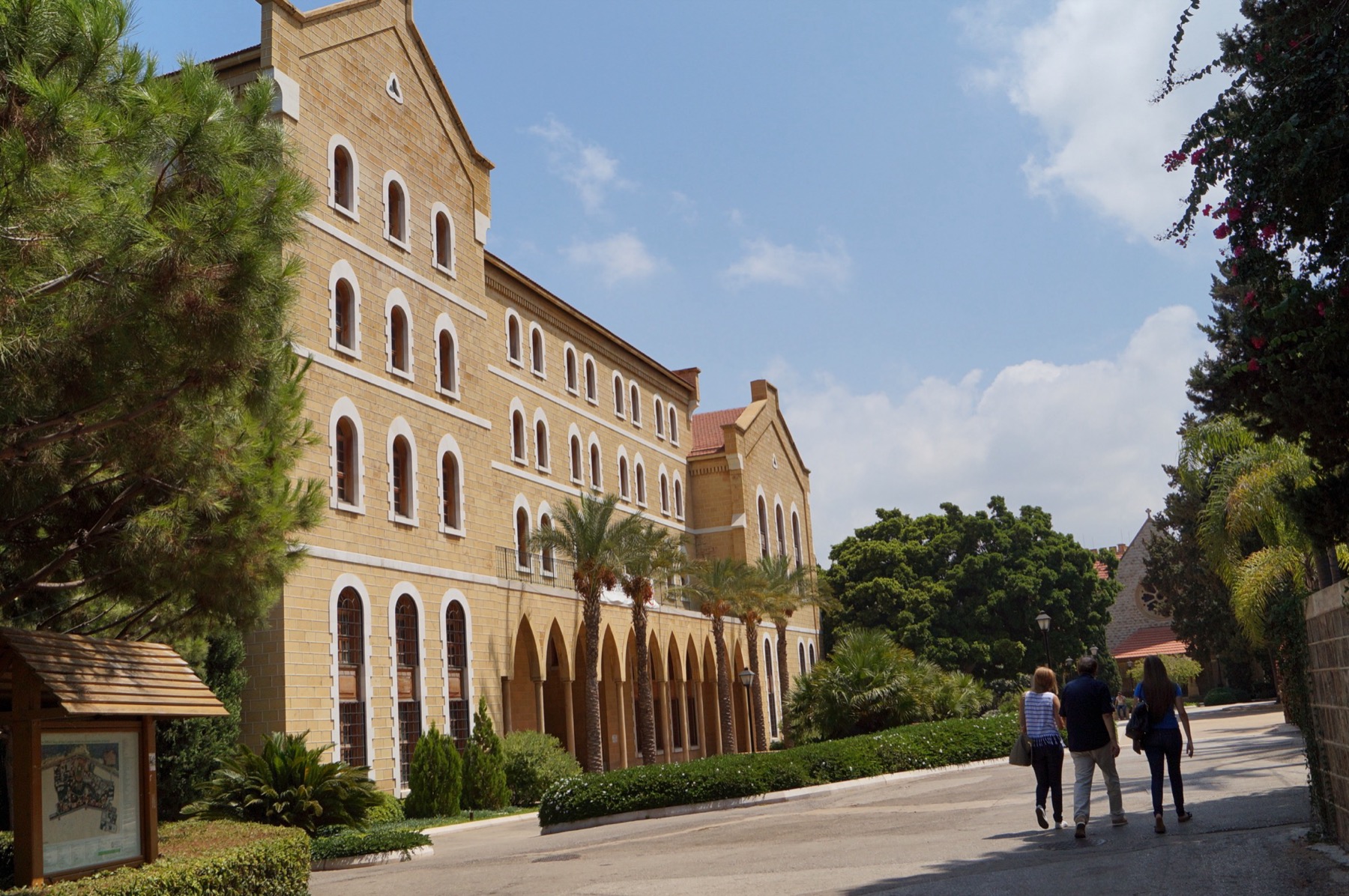 College Hall, a long sandstone building with arched windows, on the American University of Beirut campus, with palm trees and a path.