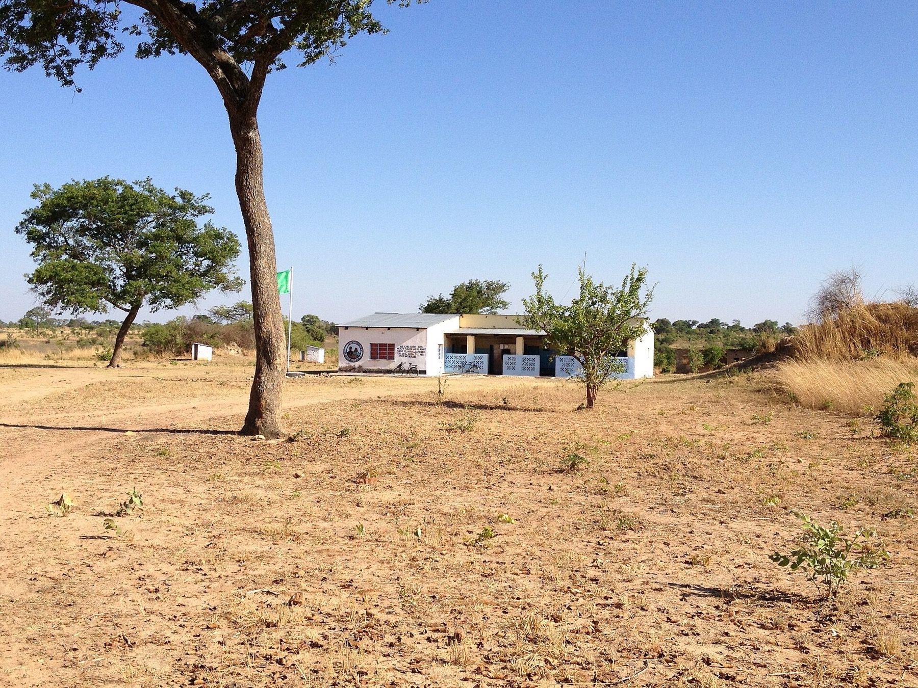 A small white single-story rural health center with a veranda and a flag pole, in dry open bush land under a blue sky.