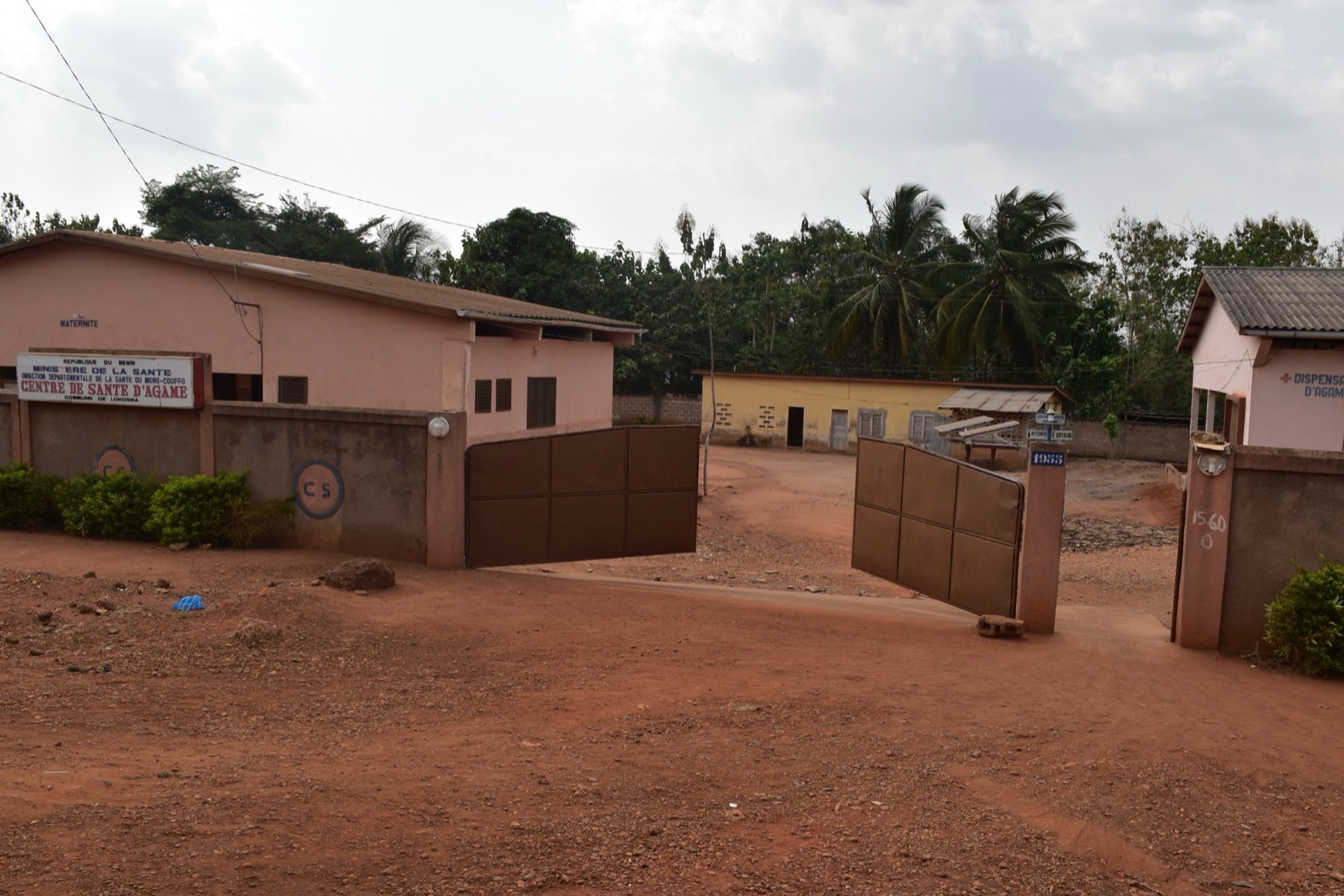 The gated entrance of a small health center in Benin: a red-earth yard, a pink building marked Maternité with a Ministry of Health sign, and a dispensary building on the right.